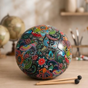 Op de afbeelding: Een bolvormige stalen tongtrommel met een levendig, veelkleurig paisley- en bloemenpatroon in blauwe, groene, oranje en rode tinten. Het instrument heeft lasergesneden tongen op het oppervlak en wordt vergezeld door twee houten hamers.