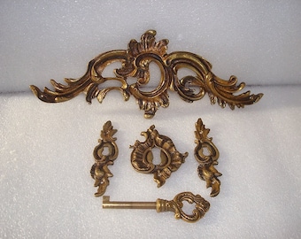 May include: A set of ornate gold-toned metal hardware, including a keyhole cover, two decorative pulls, and a key. The keyhole cover and pulls feature intricate floral designs.
