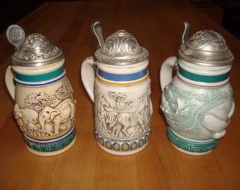 May include: Three ceramic beer steins with pewter lids and handles. Each stein features a different animal scene: elephants, zebras, and seals. The steins are decorated with blue, green, and yellow accents.