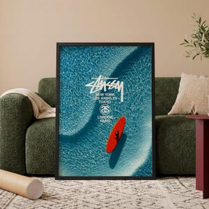 Könnte beinhalten: Ein gerahmter Kunstdruck mit einer Luftaufnahme eines leuchtend roten Surfbretts, das auf klarem, blauem Ozeanwasser treibt. Der weiße Text lautet Stussy, New York, Los Angeles, Tokyo, London, Paris, inklusive dem Stussy-Logo in der Mitte.