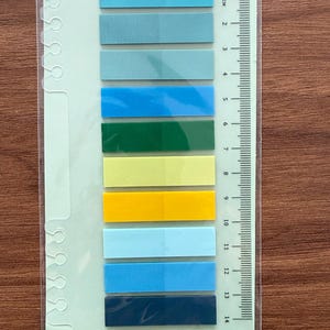 May include: A set of ten rectangular adhesive page markers in various shades of blue, green, and yellow, arranged vertically on a light green backing card that features a printed metric ruler measuring up to 14 centimeters in length.