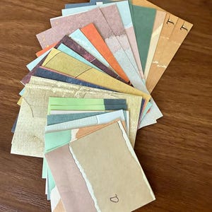 May include: A collection of square paper cards arranged in a fan shape. The cards feature various colors including muted greens, oranges, blues, and tans, with some displaying torn edges and handwritten letters like a, T, and J on their surfaces.