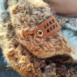 May include: A chunky knit scarf in shades of tan, brown, and cream, featuring a textured, thick yarn construction. A small, rectangular tan leather tag is attached to the fabric with a silver metal rivet, clearly embossed with the text HAND MADE.