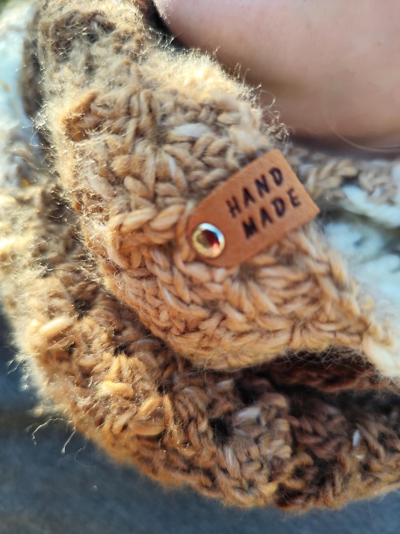 May include: A chunky knit scarf in shades of tan, brown, and cream, featuring a textured, thick yarn construction. A small, rectangular tan leather tag is attached to the fabric with a silver metal rivet, clearly embossed with the text HAND MADE.