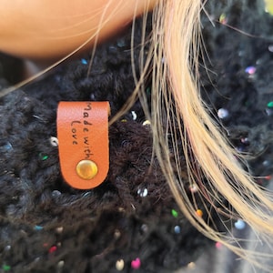 May include: A black crocheted scarf featuring embedded iridescent sequins that catch the light. A tan leather tag is attached to the fabric with a brass-colored metal rivet, featuring the text "Made with Love" embossed in black lettering.