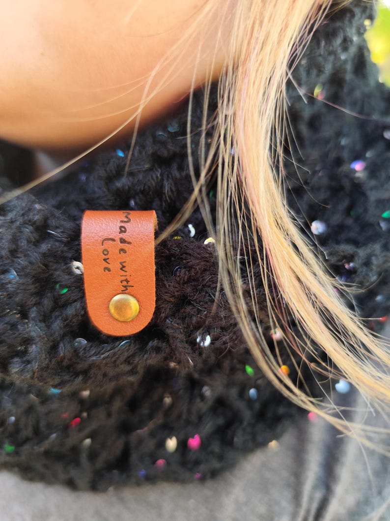 May include: A black crocheted scarf featuring embedded iridescent sequins that catch the light. A tan leather tag is attached to the fabric with a brass-colored metal rivet, featuring the text "Made with Love" embossed in black lettering.