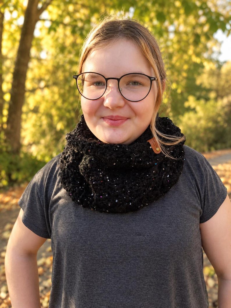 May include: A black crocheted infinity scarf featuring a textured knit pattern with subtle integrated metallic sparkles and a small brown rectangular leather tag attached to the side, worn over a plain dark gray short-sleeved t-shirt.