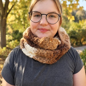 May include: A chunky crochet infinity scarf featuring a gradient of brown, tan, and cream yarn colors. The scarf has a textured, open-stitch pattern and includes a small, rectangular brown leather tag with the text "HAND MADE" in gold lettering.