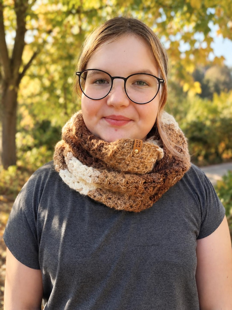 May include: A chunky crochet infinity scarf featuring a gradient of brown, tan, and cream yarn colors. The scarf has a textured, open-stitch pattern and includes a small, rectangular brown leather tag with the text "HAND MADE" in gold lettering.
