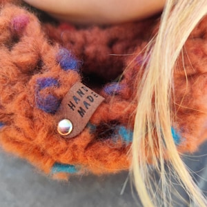May include: A chunky knit cowl in a burnt orange yarn with flecks of blue and purple. A small brown leather tag is attached to the side with a silver metal rivet, featuring the text HAND MADE embossed in black capital letters.