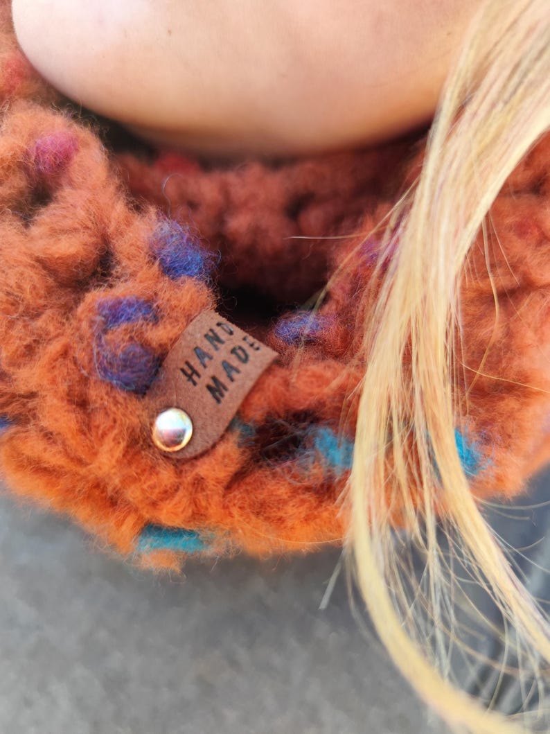 May include: A chunky knit cowl in a burnt orange yarn with flecks of blue and purple. A small brown leather tag is attached to the side with a silver metal rivet, featuring the text HAND MADE embossed in black capital letters.