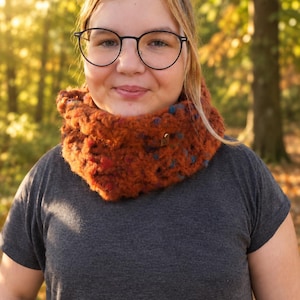 May include: A chunky, hand-knit cowl scarf in a deep burnt orange color with flecks of blue and red throughout. The scarf features a thick, textured yarn construction and a small, metallic, heart-shaped charm attached to the front side.
