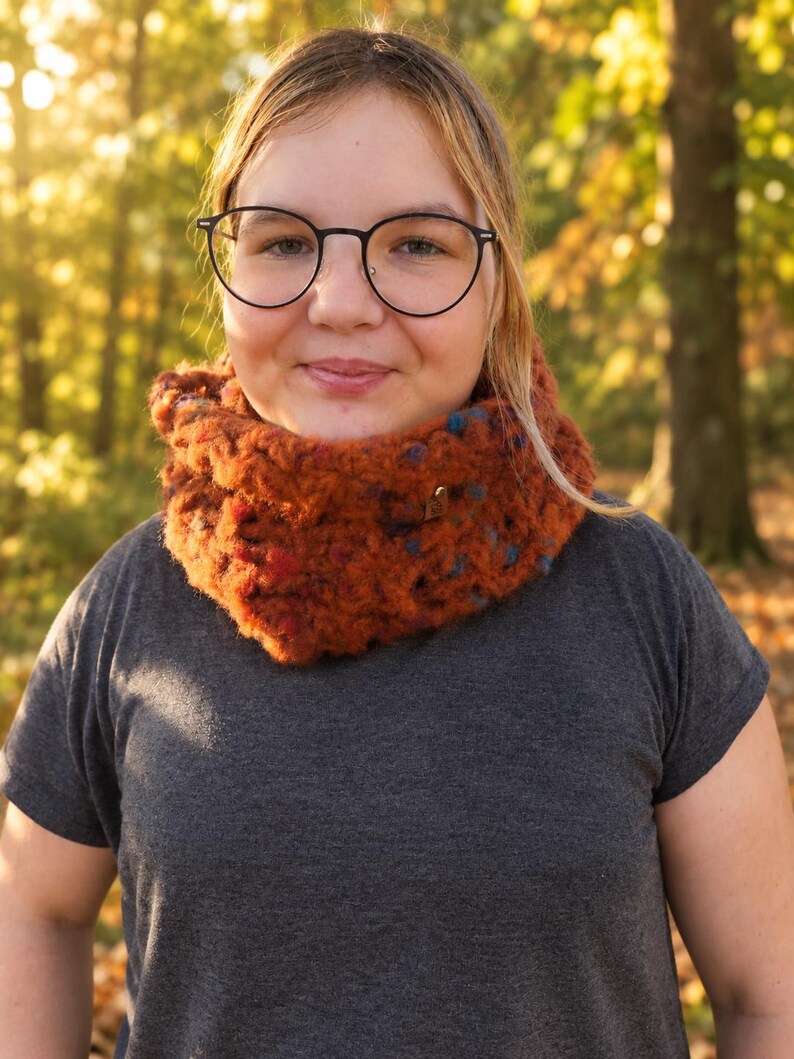 May include: A chunky, hand-knit cowl scarf in a deep burnt orange color with flecks of blue and red throughout. The scarf features a thick, textured yarn construction and a small, metallic, heart-shaped charm attached to the front side.