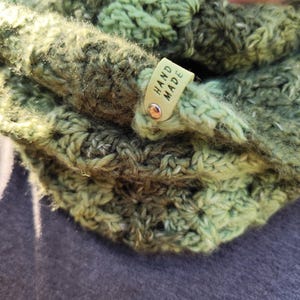 May include: A chunky crochet cowl in variegated shades of forest green, olive, and lime. The textured yarn features a repeating stitch pattern and includes a small, light-colored rectangular tag attached with a metal rivet, clearly displaying the text HAND MADE.