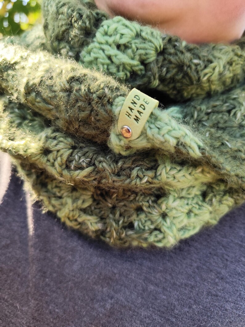 May include: A chunky crochet cowl in variegated shades of forest green, olive, and lime. The textured yarn features a repeating stitch pattern and includes a small, light-colored rectangular tag attached with a metal rivet, clearly displaying the text HAND MADE.