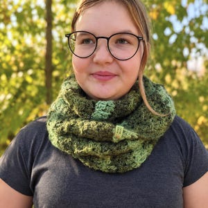 May include: A chunky crochet cowl scarf featuring a variegated blend of dark forest green, olive, and lime green yarn. The textured, circular neck warmer includes a small, rectangular light green tag with the text "HAND MADE" attached to the side.