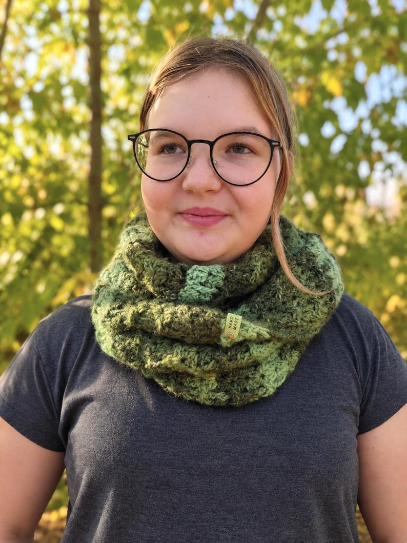 May include: A chunky crochet cowl scarf featuring a variegated blend of dark forest green, olive, and lime green yarn. The textured, circular neck warmer includes a small, rectangular light green tag with the text "HAND MADE" attached to the side.