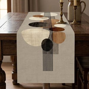 Può includere: Un runner da tavola rettangolare in tessuto beige con un motivo stampato di cerchi sovrapposti nei toni del nero, marrone e crema, accentuato da linee verticali nere che si estendono lungo il centro della superficie tessile.