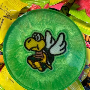 May include: A circular, translucent green resin coaster with a shimmering, glittery finish. Embedded in the center is a small, embroidered patch featuring a yellow, winged turtle character with a brown shell and white wings, outlined in black.