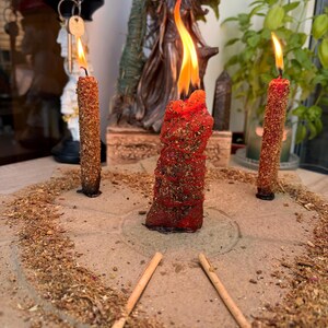 May include: Three red candles coated in dried herbs and flowers are burning on a circular stone base. The central candle is thicker and taller than the two thinner candles flanking it. Dried botanical material is scattered across the stone surface.