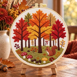 May include: A circular embroidery hoop featuring a stitched autumn forest scene with red, orange, and yellow trees, a brown squirrel, and various mushrooms on a green ground, all set within a light-coloured wooden frame on a small stand.