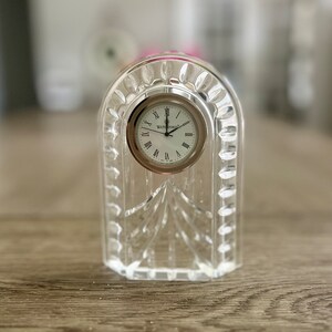 May include: A clear crystal clock with a silver face and Roman numeral hour markers. The clock is made by Waterford.