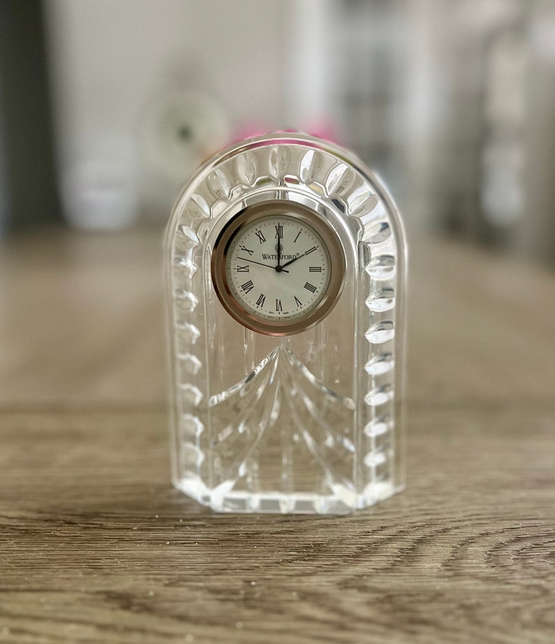 May include: A clear crystal clock with a silver face and Roman numeral hour markers. The clock is made by Waterford.