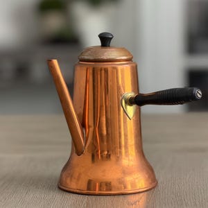 May include: A copper-colored vintage tea kettle with a black handle and lid. The kettle has a long spout and a wooden handle. The kettle is sitting on a wooden surface.