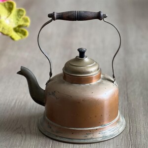 May include: A vintage copper teapot with a wooden handle and a brass lid. The teapot has a worn, aged patina and is sitting on a wooden surface.