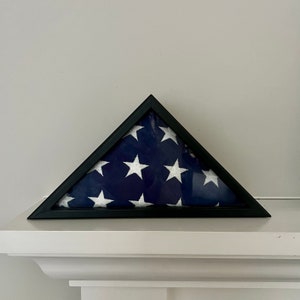 Könnte beinhalten: Ein schwarzer dreieckiger Rahmen zeigt eine gefaltete amerikanische Flagge. Die Flagge wird traditionell präsentiert, mit den Sternen in der oberen linken Ecke und den Streifen horizontal.