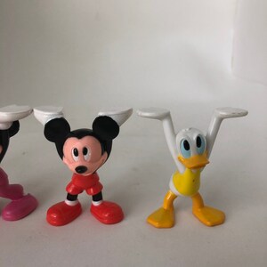 K&ouml;nnte beinhalten: Drei Plastikfiguren von Mickey Mouse, Minnie Mouse und Donald Duck. Die Figuren stehen alle mit erhobenen Armen &uuml;ber dem Kopf. Mickey Mouse tr&auml;gt eine rote Hose, Minnie Mouse ein rosa Kleid und Donald Duck ein gelbes Hemd.