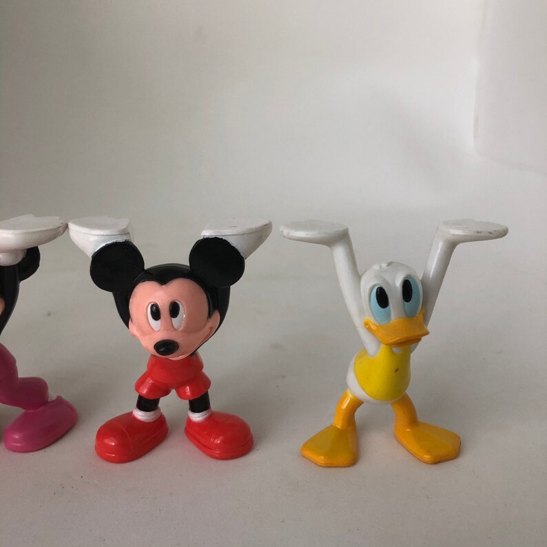K&ouml;nnte beinhalten: Drei Plastikfiguren von Mickey Mouse, Minnie Mouse und Donald Duck. Die Figuren stehen alle mit erhobenen Armen &uuml;ber dem Kopf. Mickey Mouse tr&auml;gt eine rote Hose, Minnie Mouse ein rosa Kleid und Donald Duck ein gelbes Hemd.