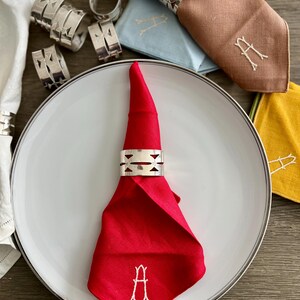 May include: A red linen napkin with a white embroidered "A" is folded and placed on a white plate with a silver rim. Silver napkin rings and other colorful napkins are also visible, suggesting a dining setting.
