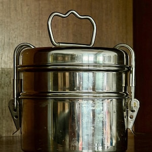 Op de afbeelding: Een roestvrijstalen tiffin-drager met twee niveaus. Het heeft een handvat en zijsluitingen. Het metaal heeft een reflecterend oppervlak. De tiffin is ontworpen voor het vervoeren van voedsel.