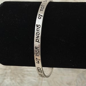 May include: A silver-toned metal bangle bracelet featuring engraved black lettering. The visible text on the outer surface of the bracelet reads, "to guard you in all you". The bracelet has a smooth, polished finish and a thin, circular band design.