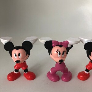 K&ouml;nnte beinhalten: Drei kleine Plastikfiguren von Mickey Mouse und Minnie Mouse. Mickey Mouse tr&auml;gt eine rote Hose und schwarze Schuhe. Minnie Mouse tr&auml;gt eine rosa Hose und rosa Schuhe. Die Figuren stehen alle aufrecht.