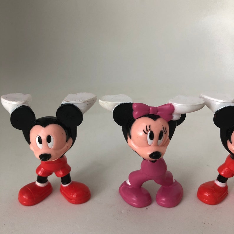 K&ouml;nnte beinhalten: Drei kleine Plastikfiguren von Mickey Mouse und Minnie Mouse. Mickey Mouse tr&auml;gt eine rote Hose und schwarze Schuhe. Minnie Mouse tr&auml;gt eine rosa Hose und rosa Schuhe. Die Figuren stehen alle aufrecht.