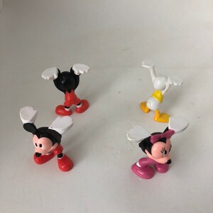 K&ouml;nnte beinhalten: Vier kleine Plastikfiguren von Disney-Charakteren. Mickey Mouse, Minnie Mouse, Donald Duck und Goofy sind alle in verschiedenen Posen mit erhobenen Armen.