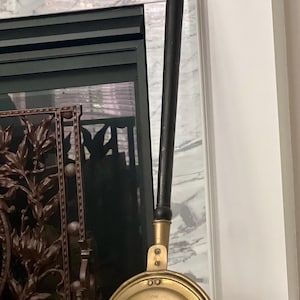 May include: A brass fireplace tool with a black wooden handle. The tool has a round, decorative head with an intricate design.