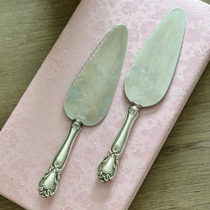 May include: Two silver cake servers with ornate handles rest on a pink floral patterned surface. One server has a serrated edge. The servers have a classic, elegant design, perfect for serving cakes and pastries.