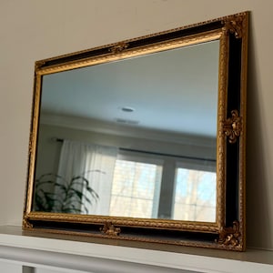 May include: Rectangular mirror with a black and gold ornate frame. The frame has intricate gold detailing and a black inner border. The mirror is displayed on a white mantelpiece, reflecting a room with a window and curtains.
