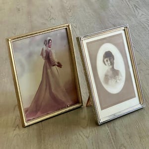 May include: Two vintage picture frames with gold and silver metal finishes. One frame holds a color photo of a woman in a long dress, the other a black and white portrait. Both frames are displayed on a wooden surface.
