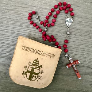 Könnte beinhalten: Ein Rosenkranz mit roten Perlen und einem silbernen Kruzifix. Die Perlen sind wie Rosen geformt. Ein beiges Täschchen mit dem Text "TERTIUM MILLENNIUM" und einem religiösen Emblem liegt neben dem Rosenkranz.