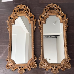 Puede incluir: Dos espejos dorados ornamentados, con copetes arqueados y volutas decorativas. Los espejos rectangulares se exhiben sobre una superficie de madera oscura, reflejando el interior de la habitación. Los marcos presentan detalles intrincados, agregando elegancia.