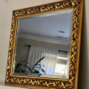 May include: Square gold mirror with ornate, detailed frame. The frame features a repeating leaf and vine pattern. The mirror reflects a room with a white curtain and a green plant. The mirror is resting on a white surface.