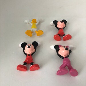 K&ouml;nnte beinhalten: Vier kleine Plastikfiguren: eine gelbe Ente, eine rot-schwarze Mickey Maus, eine rot-schwarze Minnie Maus und eine pink-schwarze Minnie Maus. Alle Figuren stehen mit erhobenen Armen &uuml;ber dem Kopf.