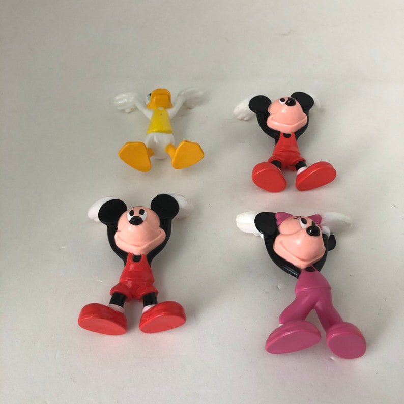 K&ouml;nnte beinhalten: Vier kleine Plastikfiguren: eine gelbe Ente, eine rot-schwarze Mickey Maus, eine rot-schwarze Minnie Maus und eine pink-schwarze Minnie Maus. Alle Figuren stehen mit erhobenen Armen &uuml;ber dem Kopf.