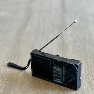May include: A black FM/AM full-range receiver radio with a retractable aerial and a wrist strap. The radio has a digital display and several buttons. The text "FULLRANGE" and "FM/AM RECEIVER" are visible on the front.