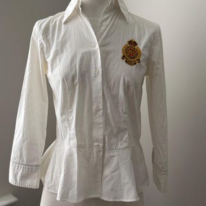 Peut inclure: Une chemise blanche boutonnée avec une taille péplum et des manches longues. La chemise présente un écusson décoratif sur la poitrine avec des détails dorés, rouges et noirs. La chemise est présentée sur un mannequin.