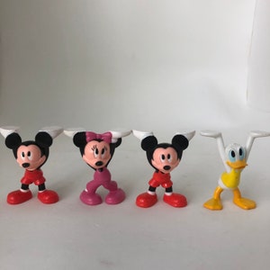 K&ouml;nnte beinhalten: Ein Set mit vier kleinen Plastikfiguren von Disney-Charakteren. Mickey Mouse, Minnie Mouse, Donald Duck und Goofy tragen alle rote, rosa, gelbe und blaue Outfits. Die Figuren sind mit erhobenen Armen &uuml;ber ihren K&ouml;pfen posiert.